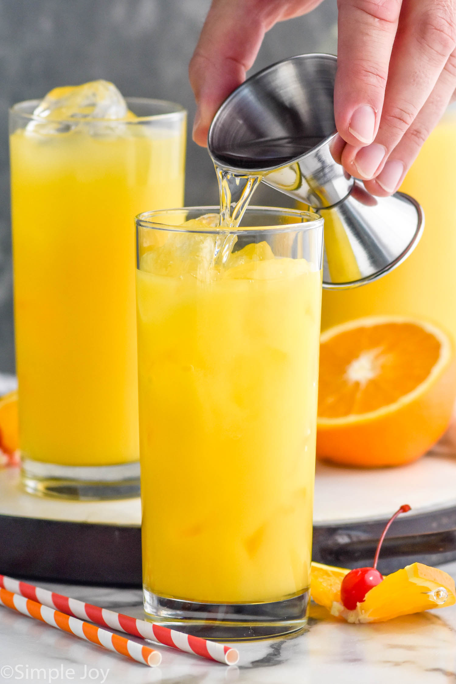 Side view of person's hand pouring ingredients into glass for Tequila Sunrise recipe. Another glass of ingredients for Tequila Sunrise on tray in background, along with fruit and straws.