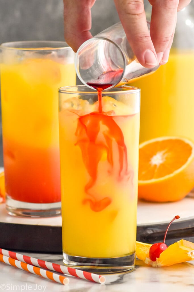 Side view of person's hand pouring grenadine into glass of ingredients for Tequila Sunrise recipe. Glasses of ingredients on tray behind and fruit and straws beside.