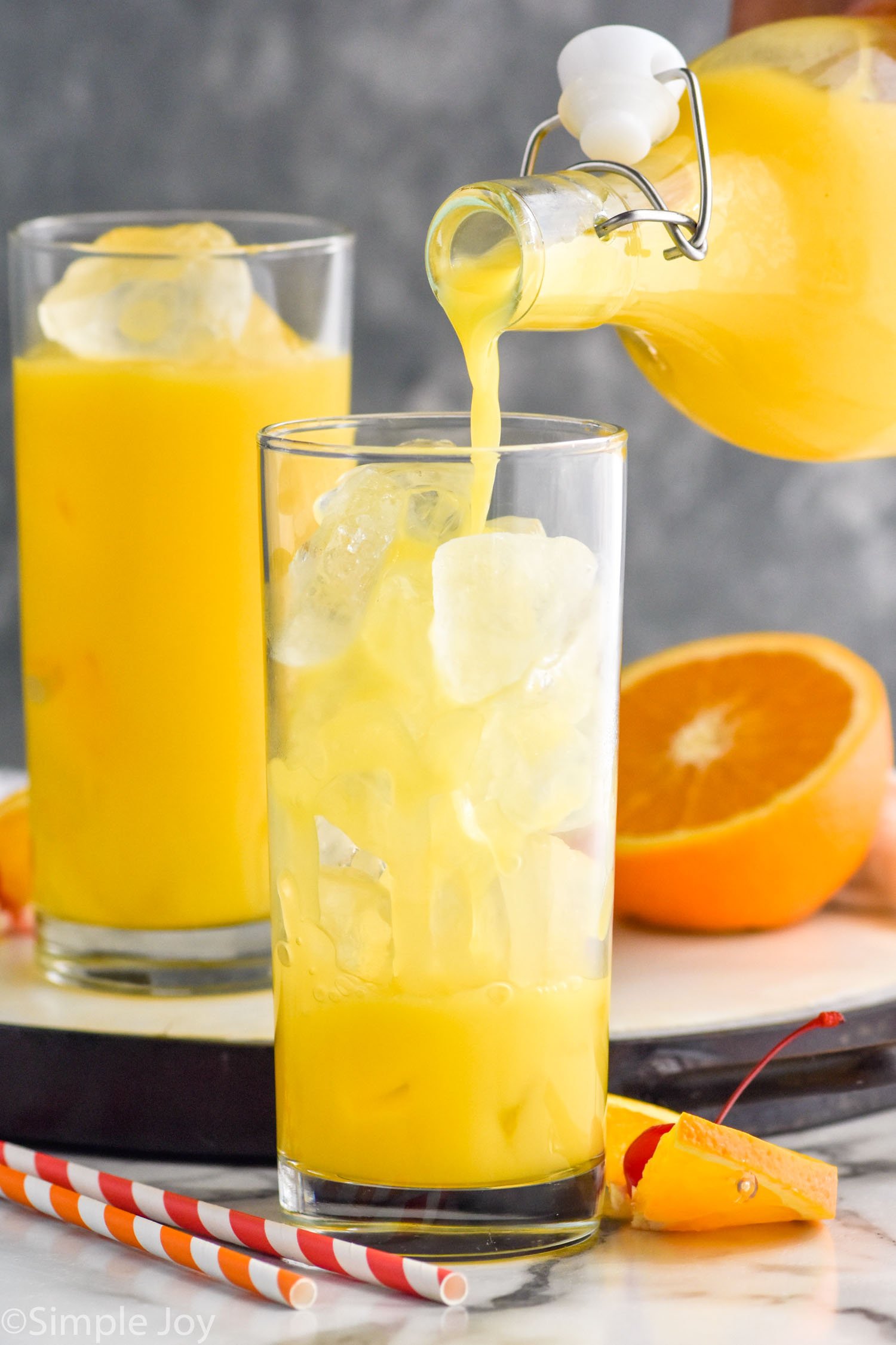Side view of orange juice being poured into glass of ice for Tequila Sunrise recipe. Another glass of juice in background and fruit on counter for garnish.