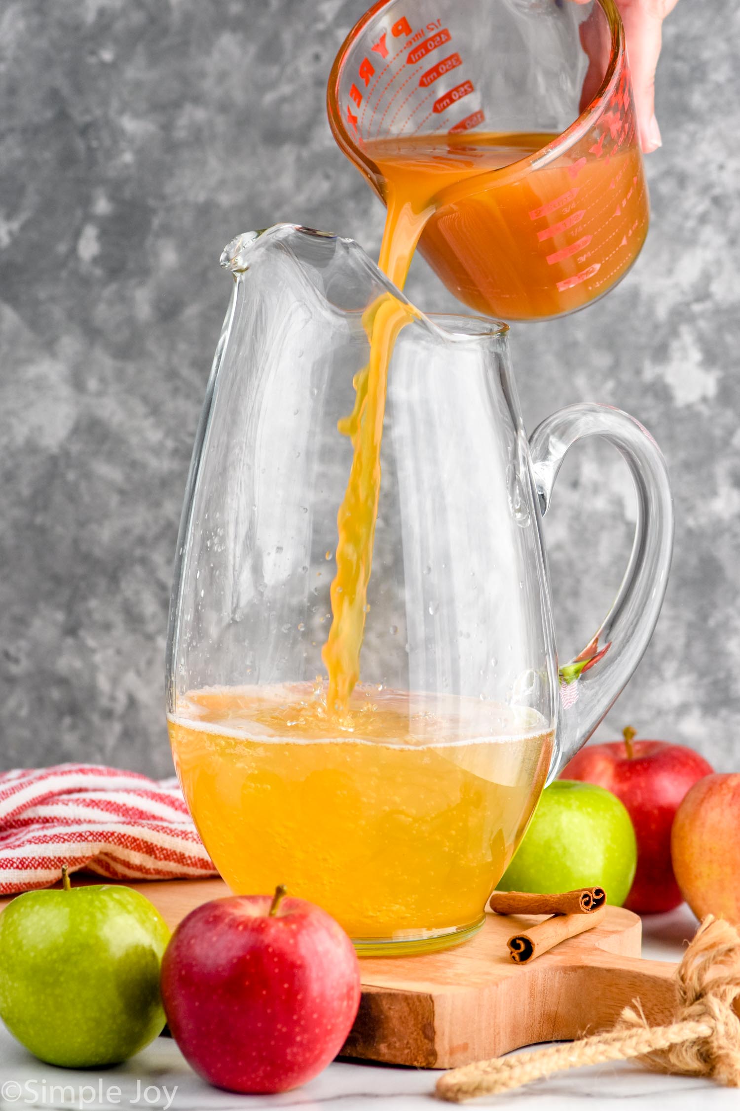 Side view of person's hand pouring ingredients into pitcher for Apple Cider Sangria recipe. Apples and cinnamon sticks beside pitcher.