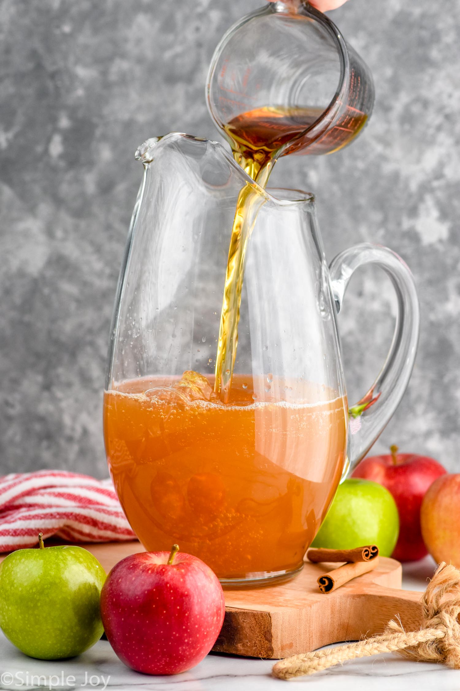 Side view of ingredients being poured into pitcher for Apple Cider Sangria recipe. Apples and cinnamon sticks beside pitcher.