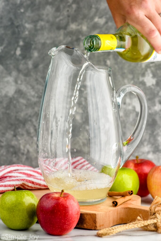 Side view of person's hand pouring bottle of wine into pitcher for Apple Cider Sangria recipe. apples and cinnamon sticks beside pitcher