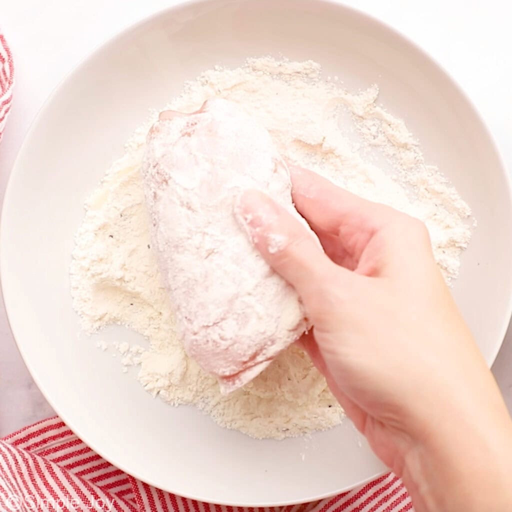 Person's hand holding piece of chicken cordon bleu into bowl of flour mixture to make chicken cordon bleu
