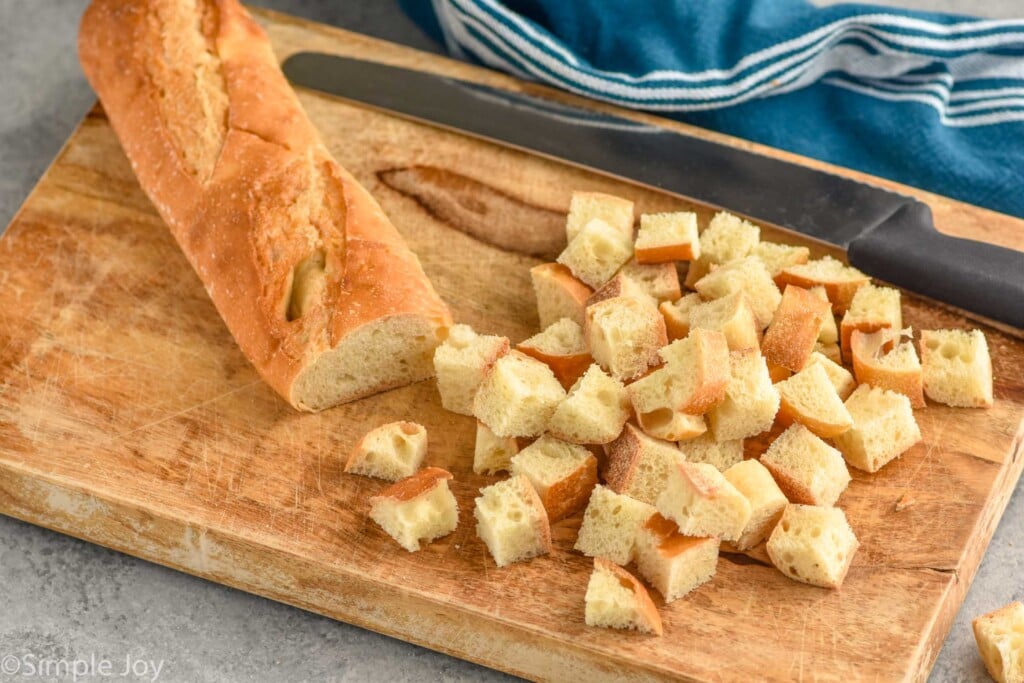 Photo of a loaf of bread partially diced into cubes for Croutons recipe on cutting board with knife.