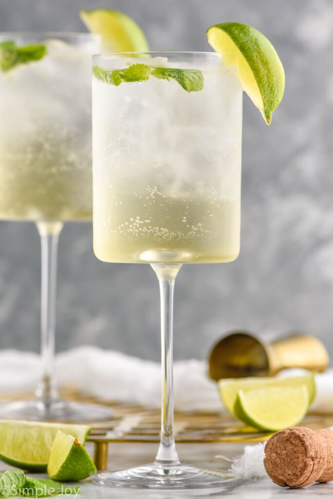 Side view of two wine glasses of Hugo Spritz garnished with lime wedges. Cork, cocktail jigger, and extra lime wedges beside glasses