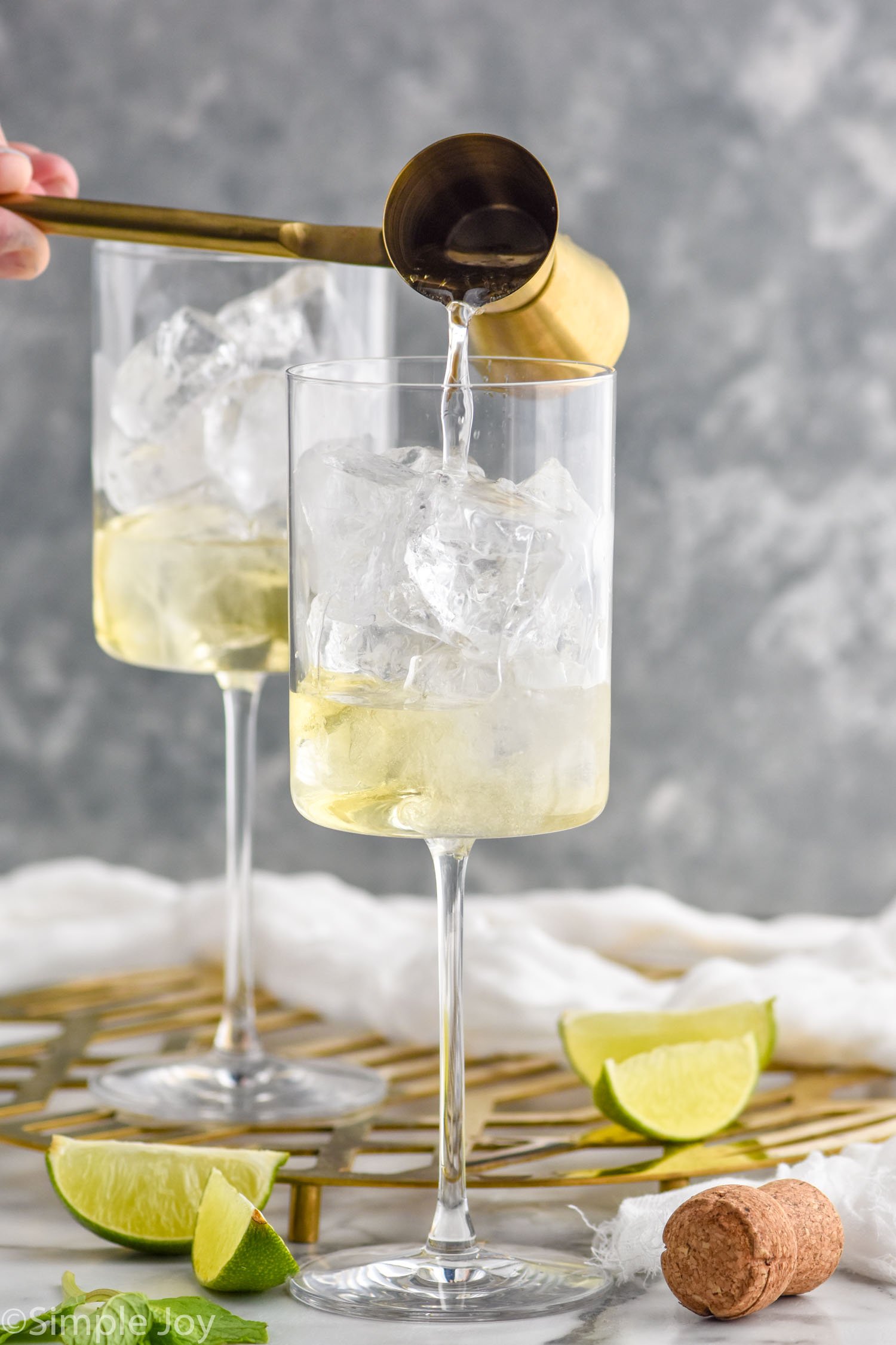 Side view of cocktail jigger pouring ingredient into wine glasses for Hugo Spritz recipe. Lime wedges on counter beside wine glasses.