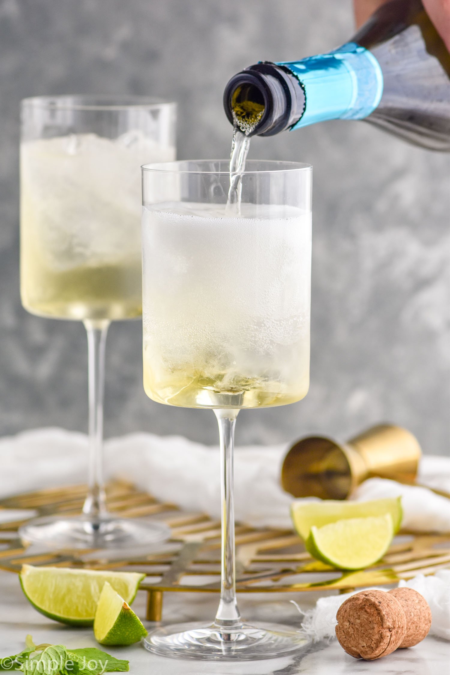 Side view of sparkling wine being poured into wine glasses for Hugo Spritz recipe. Lime wedges and cocktail jigger on counter beside glasses.