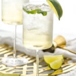 Side view of two wine glasses of Hugo Spritz garnished with lime wedges. Cork, cocktail jigger, and extra lime wedges beside glasses - 10