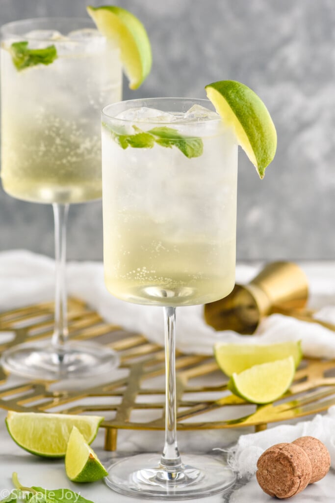 Side view of two wine glasses of Hugo Spritz garnished with lime wedges. Cork, cocktail jigger, and extra lime wedges beside glasses