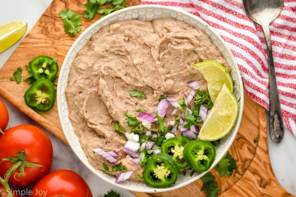 overhead of bowl of mashed pinto beans made in the instant pot. Topped with cilantro, red onion, diced jalapeno, and lime wedges. vegetables and spoon sitting beside bowl