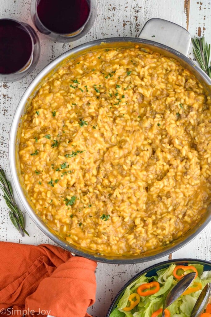 overhead of skillet of pumpkin risotto with two glasses of red wine and bowl of salad sitting beside