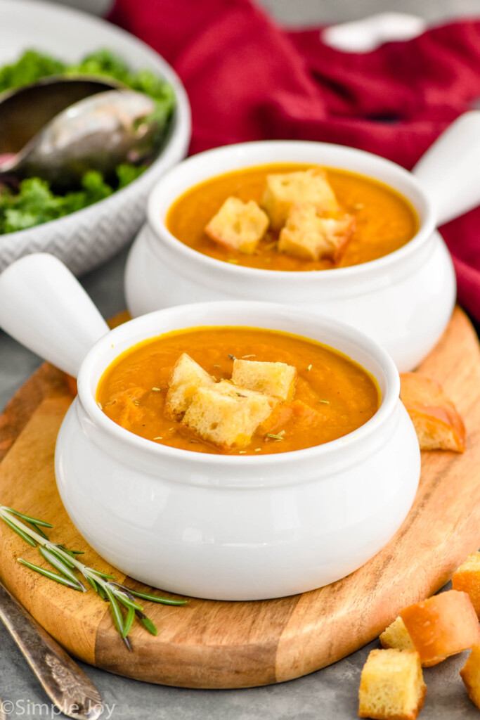 Photo of two bowls of Pumpkin Soup garnished with croutons on a wooden tray. Salad beside tray.