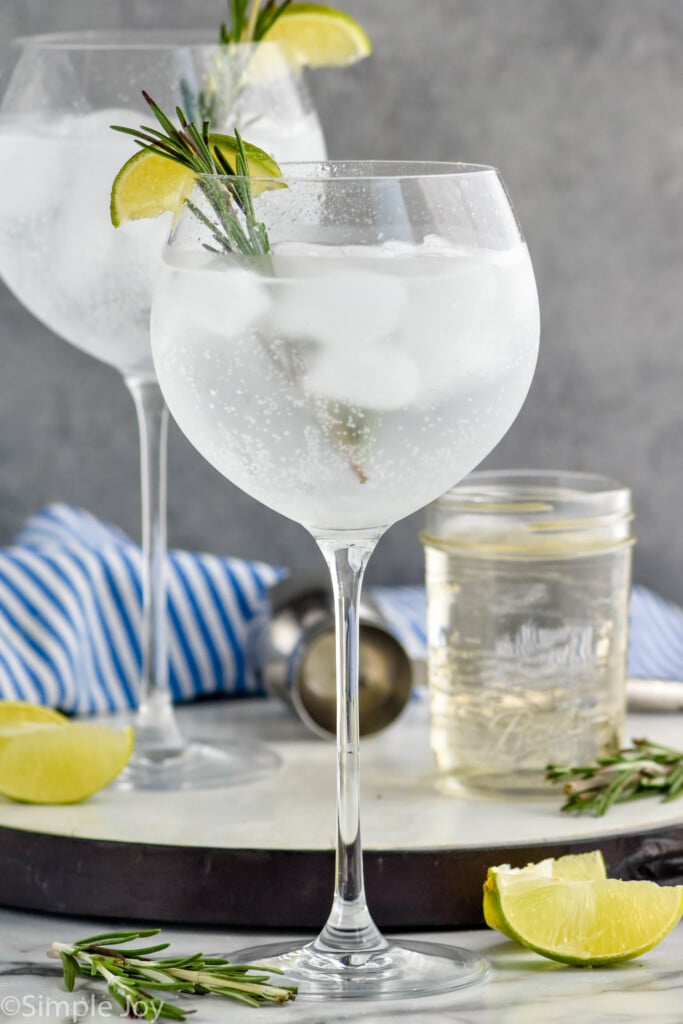 Side view of two rosemary gin and tonic cocktails beside a jar of Simple Syrup. rosemary leaves and lime wedges on counter.