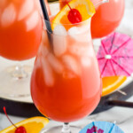 Photo of Sex on the Beach garnished with orange slice and cherry. Extra fruit and umbrellas beside glass for garnish.