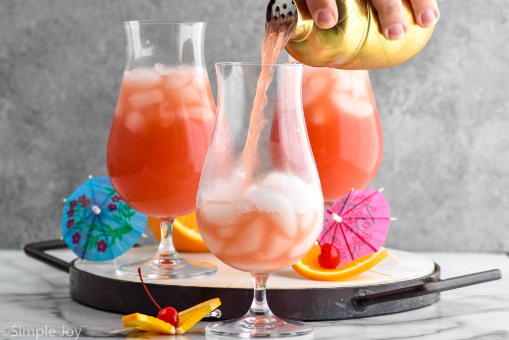 Side view of person's hand pouring cocktail shaker of ingredients into glass of ice for Sex on the Beach recipe. Orange slices, cherries, and umbrellas beside glasses for garnish.