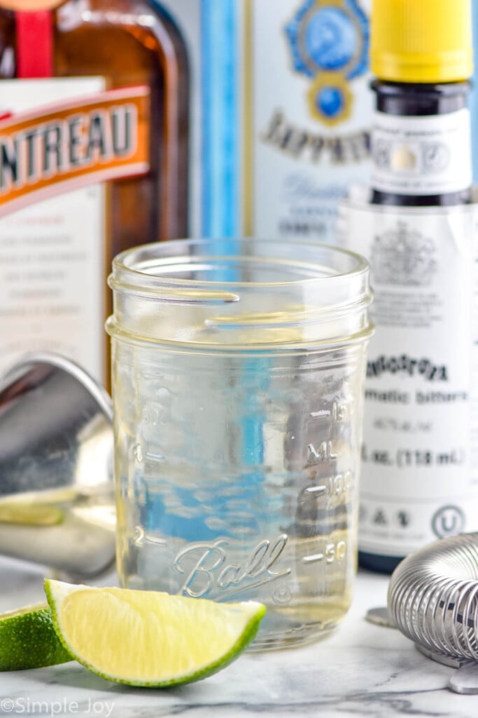 Photo of a jar of Simple Syrup recipe. Lime wedges, cocktail jigger, and bottles of liqueur next to jar.