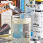 Photo of a spoon over a jar of Simple Syrup recipe. Lime wedges, cocktail jigger, and bottles of cointreau, gin, and bitters beside jar. - 11
