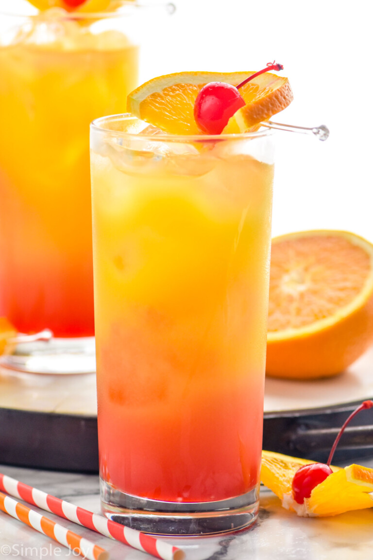 Side view of Tequila Sunrise garnished with cherry and orange slice. Another Tequila Sunrise sits on tray behind. Extra oranges for garnish and straws on counter.