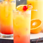 Side view of two Tequila Sunrise cocktails garnished with orange slice and cherry. Extra fruit and straws on counter.