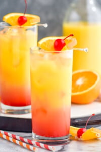 Side view of two Tequila Sunrise cocktails garnished with orange slice and cherry. Extra fruit and straws on counter.