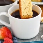 Pinterest graphic for Biscotti recipe. Image shows a person's hand dipping a Biscotti in a cup of coffee. Platter of Biscotti, fruit, and cup of coffee beside. Text says, "homemade Biscotti recipe simplejoy.com"