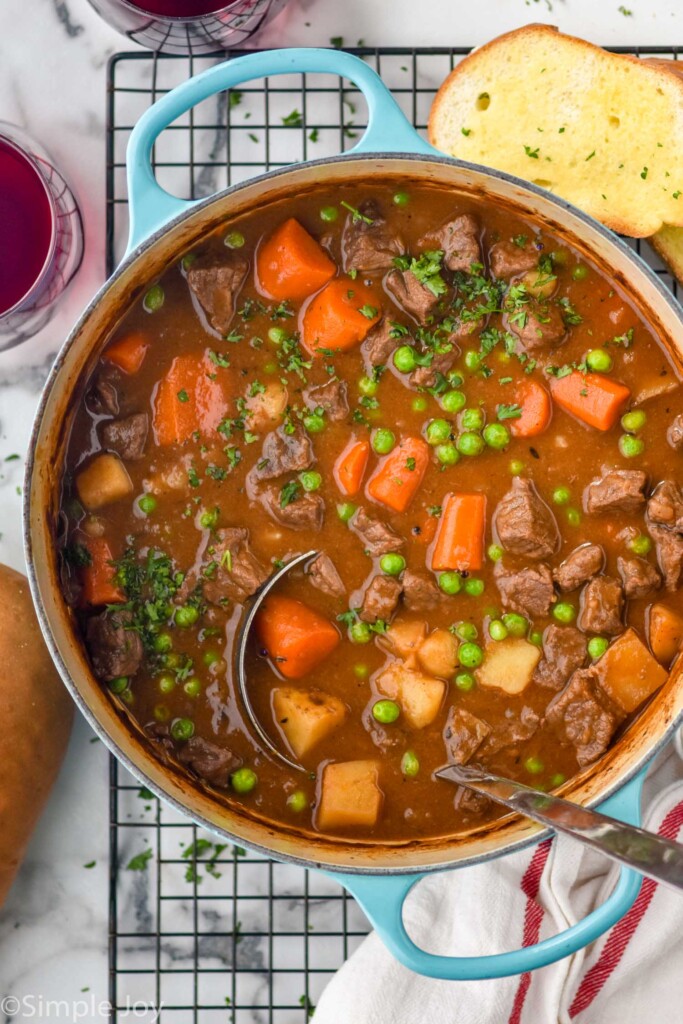 Overhead photo of a pot of Beef Stew with a ladle. Bread and glasses of wine beside.