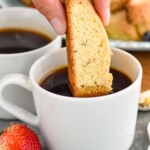 Photo of a person's hand dipping Biscotti in a cup of coffee. Cup of coffee, fruit, and plate of Biscotti beside.