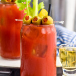 Photo of two glasses of Bloody Mary garnished with olives, celery, and pickles. Shot of beer and spear of olives beside.