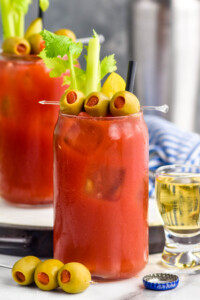 Photo of two glasses of Bloody Mary garnished with olives, celery, and pickles. Shot of beer and spear of olives beside.