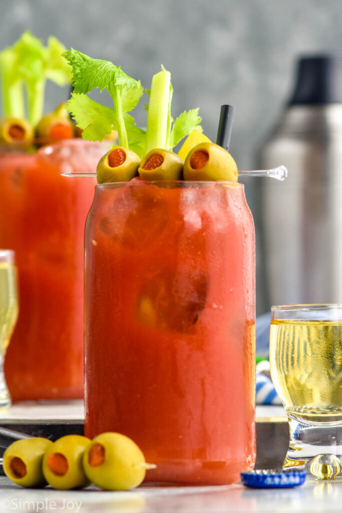 Side view of Bloody Mary cocktails garnished with olives and celery. Olives and shot of beer beside glasses.