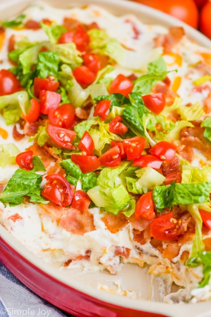up close of a ceramic baking dish with a creamy hot dip recipe topped with fresh lettuce and tomatoes, with part of the dip missing