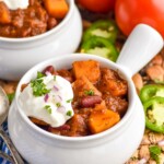 two bowls of butternut squash chili topped with sour cream. Corn chips, jalapenos, tomatoes, and spoons sitting beside