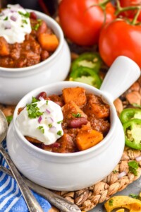 two bowls of butternut squash chili topped with sour cream. Corn chips, jalapenos, tomatoes, and spoons sitting beside