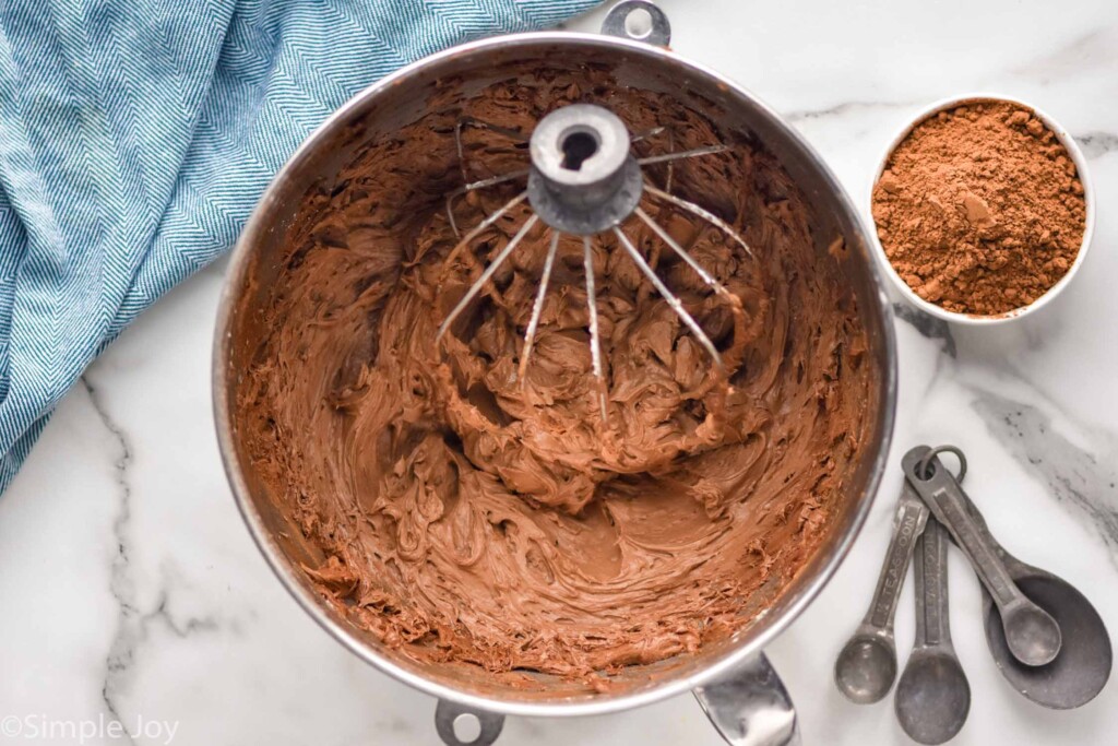 overhead of stand mixer bowl of chocolate frosting. Bowl of cocoa powder and measuring spoons sitting beside