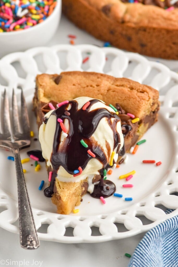 Photo of a slice of Cookie Cake served on a plate and garnished with ice cream and chocolate syrup. Fork for serving.
