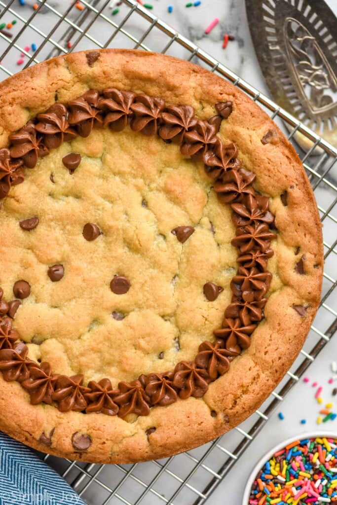 Overhead photo of Cookie Cake on a cooling rack with sprinkles and serving spatula beside