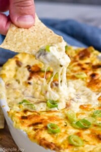 person's hand holding chip with corn dip out of a dish of hot corn dip