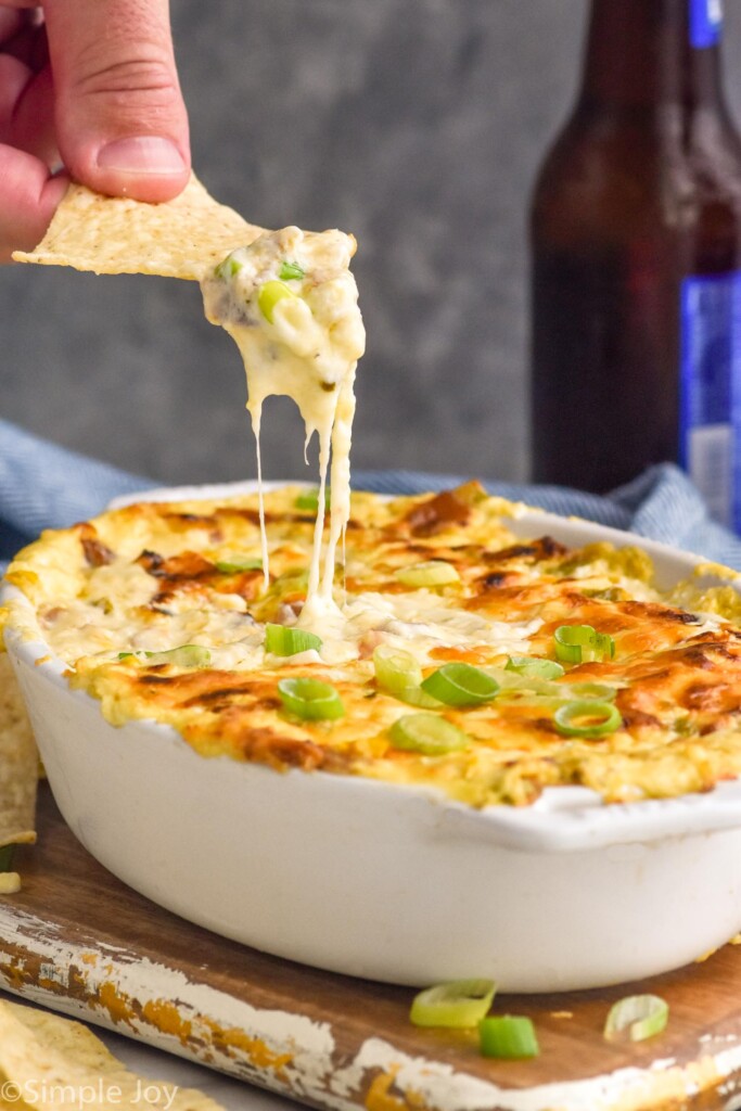 person's hand holding chip with corn dip out of a dish of hot corn dip