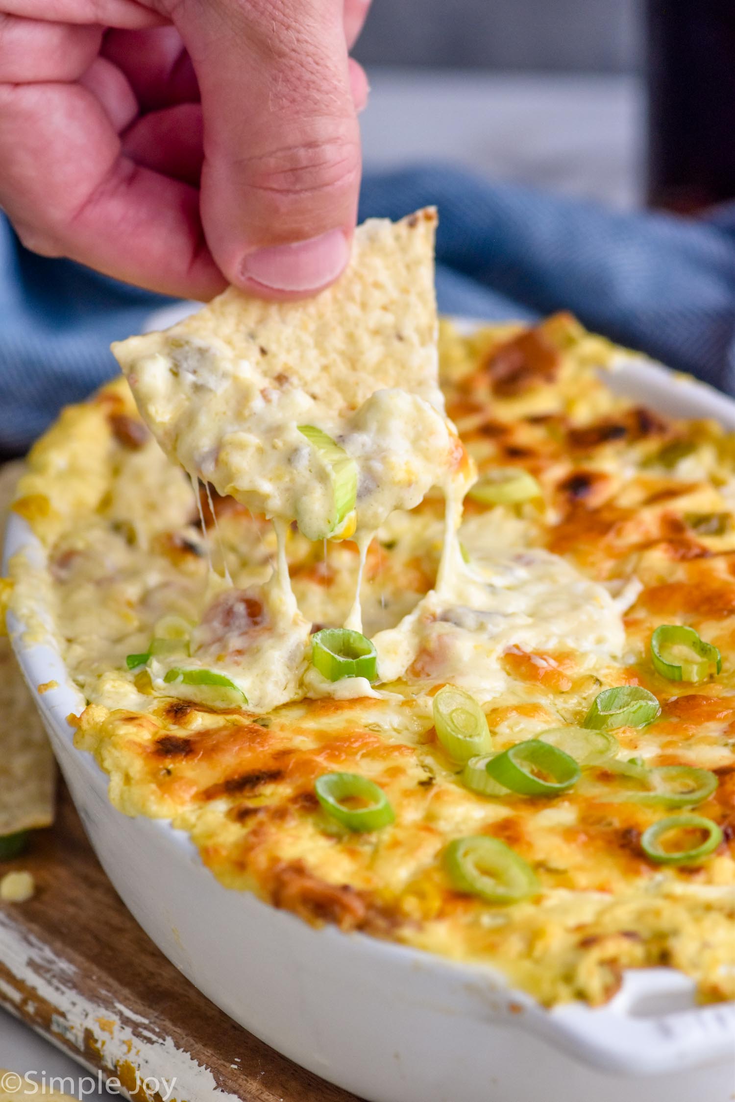 person's hand holding chip with corn dip out of a dish of hot corn dip