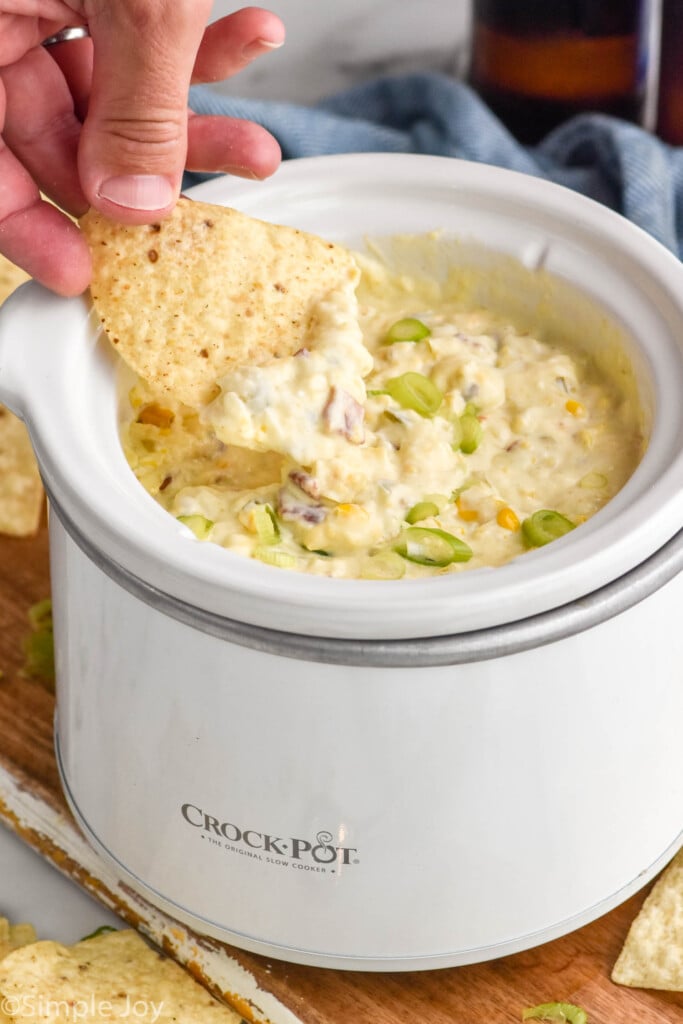 person's hand holding chip with corn dip out of small crockpot of hot corn dip