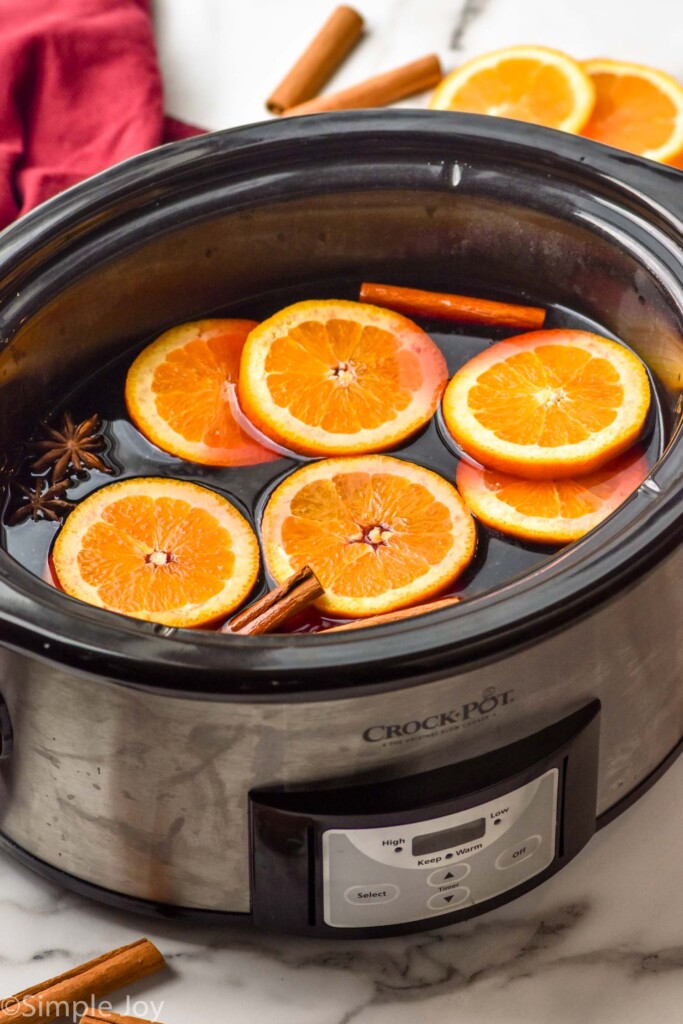 Photo of a crock pot of ingredients for Slow Cooker Mulled Wine recipe with cinnamon sticks and orange slices beside.