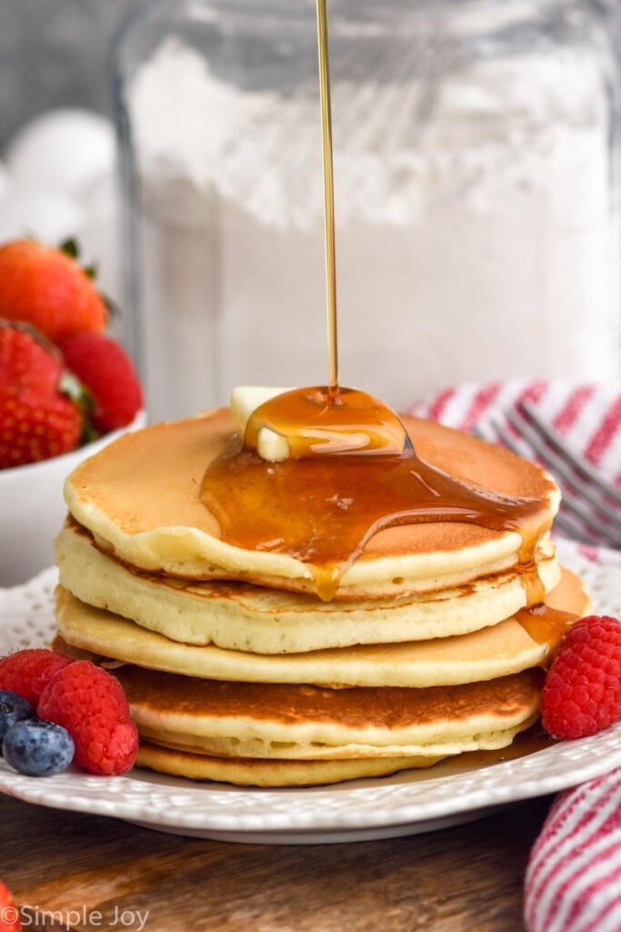 Plate of pancakes with butter and maple syrup with fresh berries. Container of pancake mix sitting in background