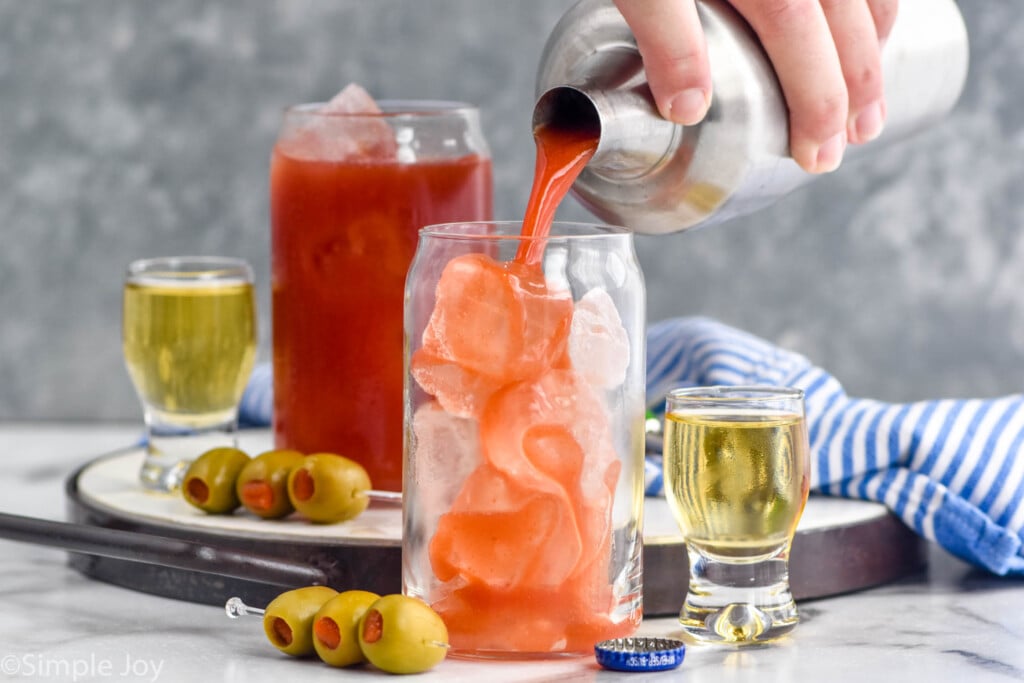 Side view of person's hand pouring cocktail shaker of Bloody Mary cocktail into a glass of ice. Another glass of Bloody Mary behind. Shots of beer and spears of olives beside.