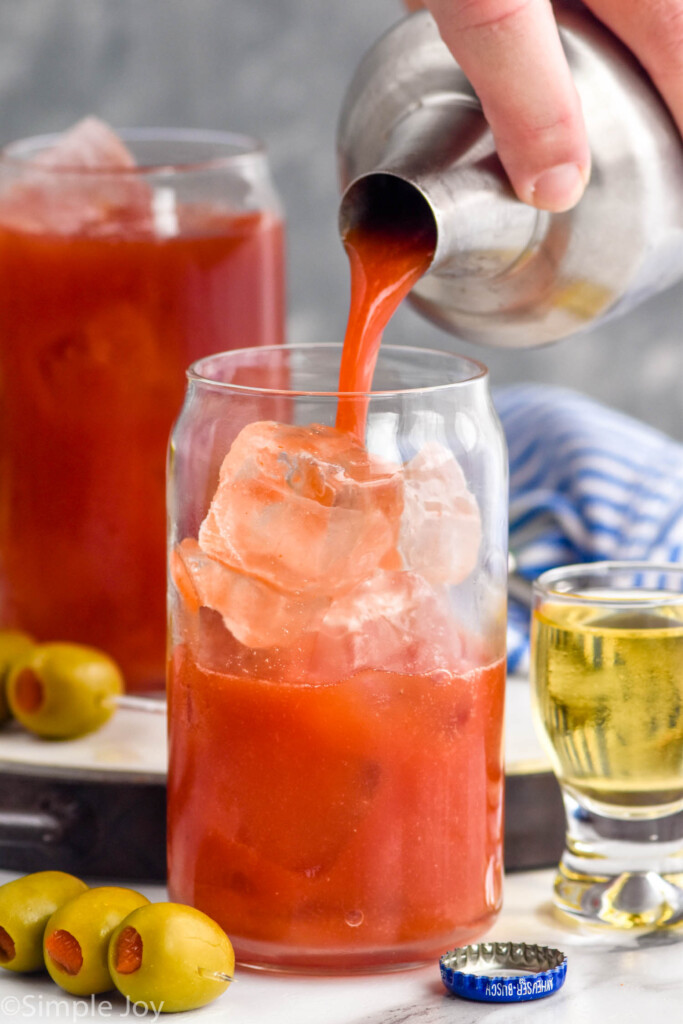Photo of person's hand pouring Bloody Mary into a glass of ice. Shot of beer and spears of olives