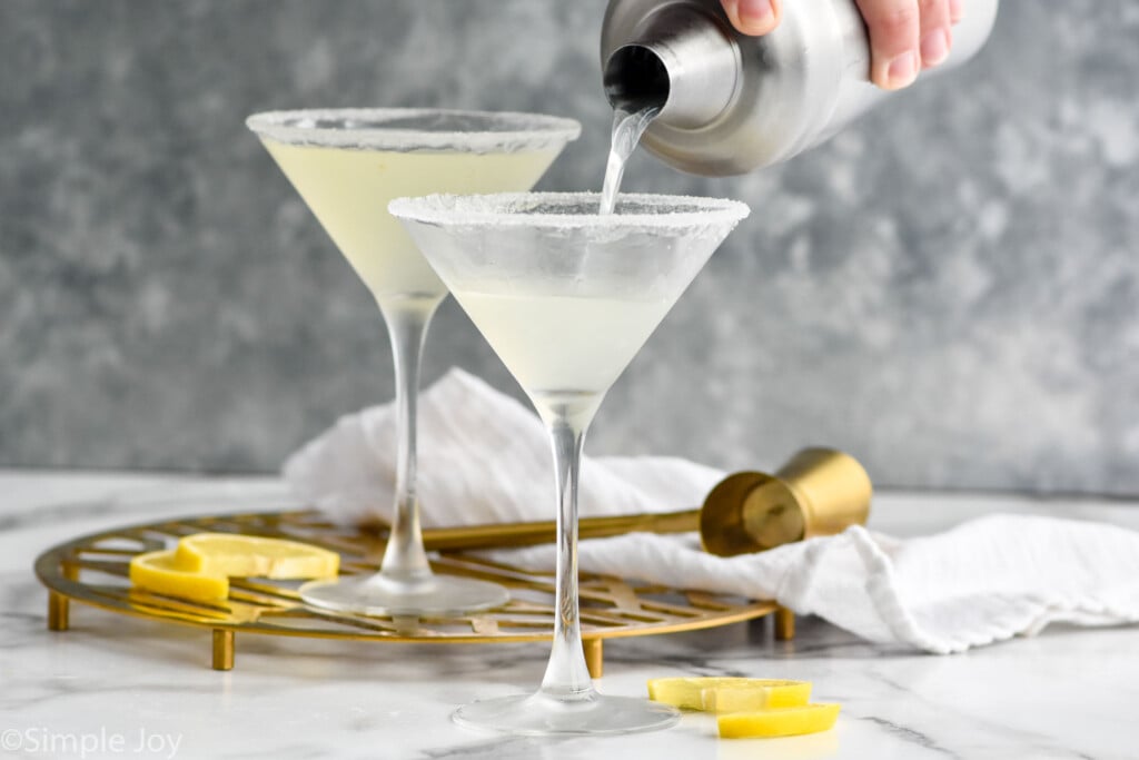 Side view of person's hand pouring martini shaker of Lemon Drop Martini recipe into martini glasses with sugar rims. Lemon slices and cocktail jigger beside glasses.