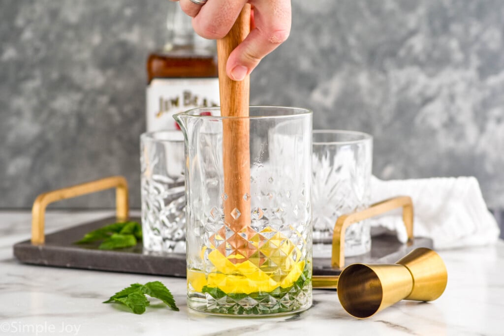 Side view of person's hand muddling ingredients in cocktail shaker for Whiskey Smash recipe. Glass tumblers, bottle of bourbon, cocktail jigger, and mint leaves beside.