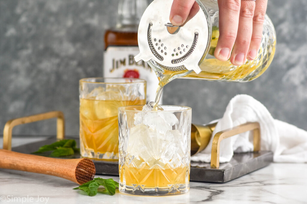 Side view of person's hand pouring cocktail shaker of ingredients into tumbler of ice for Whiskey Smash recipe. Muddler, cocktail jigger, bottle of bourbon, and another tumbler of Whiskey Smash recipe beside.