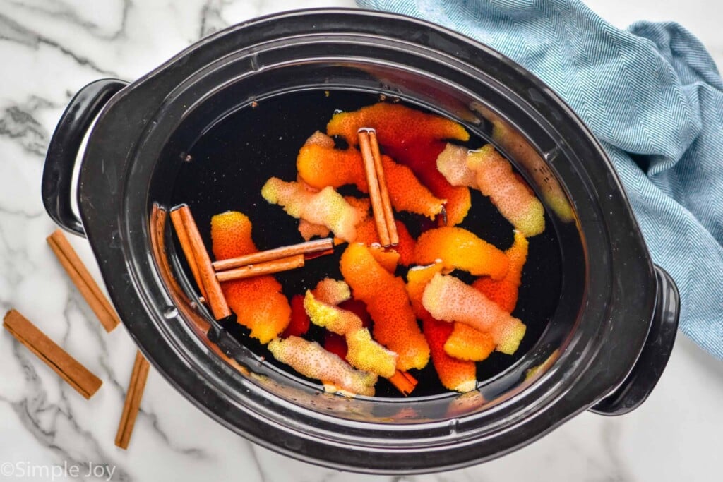 Overhead photo of crock pot of ingredients for Slow Cooker Mulled Wine recipe with cinnamon sticks and a towel beside.