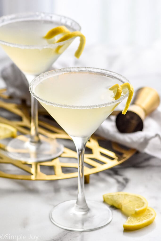 Photo of two Lemon Drop Martini cocktails garnished with twist of lemon. Lemon slices and cocktail jigger beside.
