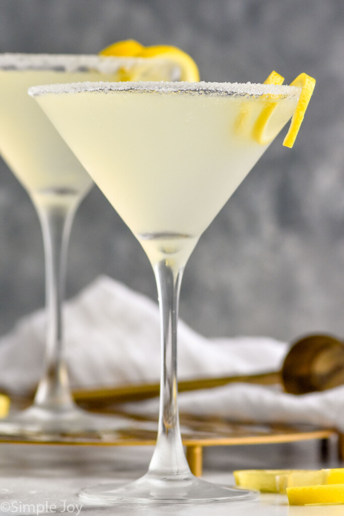 Side view of two Lemon Drop Martini cocktails with lemon wedges and cocktail jigger beside.